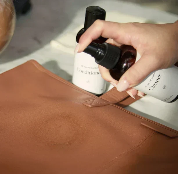 A hand spraying leather cleaner onto a tan leather handbag